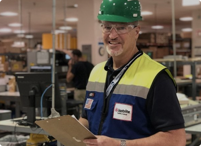 Justrite Safety Group worker in a hard hat holding a clipboard on the warehouse floor